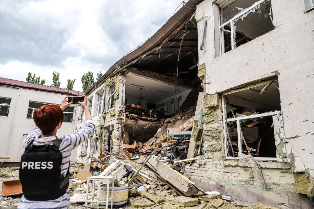 Photo of a journalist wearing a press vest taking photos of rubble of a building that was destroyed during the war. 