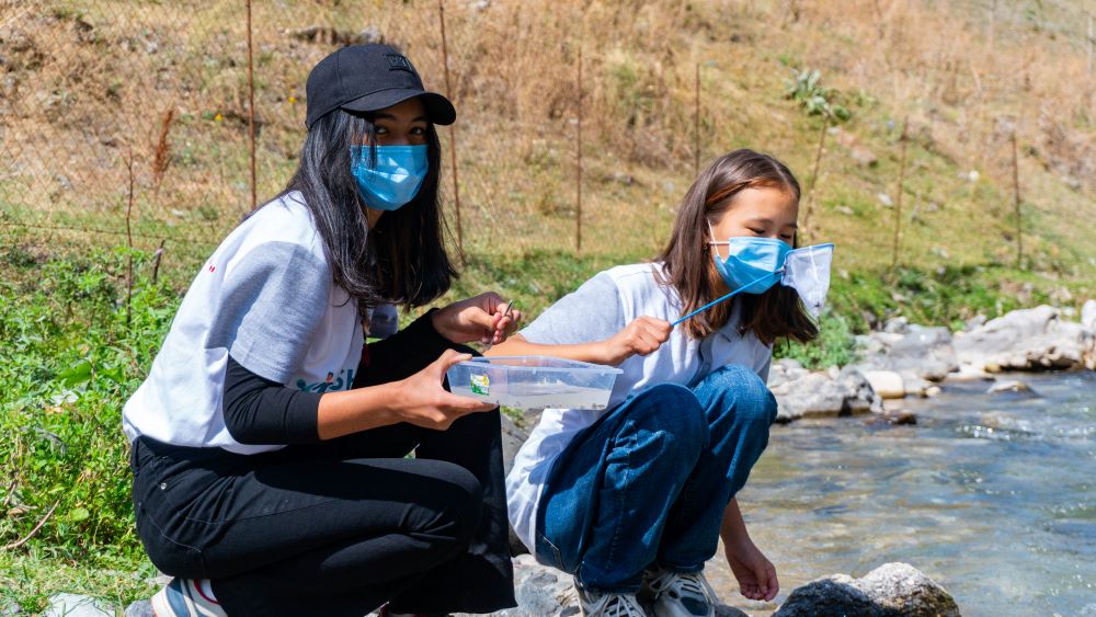 Photo of two young girls squatting by the side of a river collecting samples from the water. The girls are wearing masks.