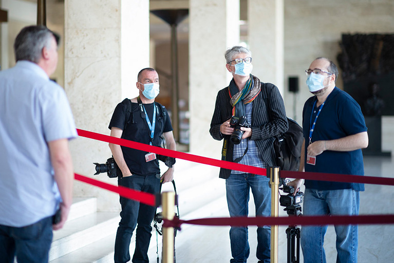 Three journalists wearing masks. They are standing on one side of a red tape barrier and talking to a person on the other side.