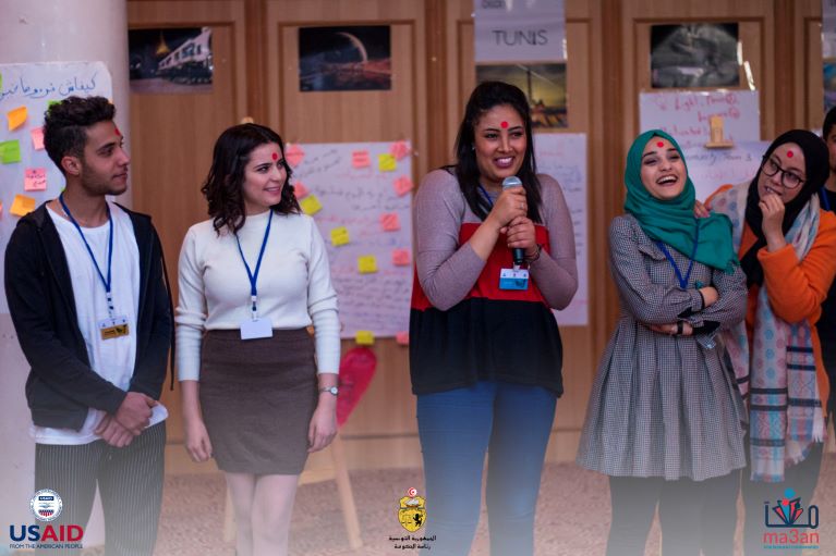 One participant speaking into a microphone with 4 others around her smiling.