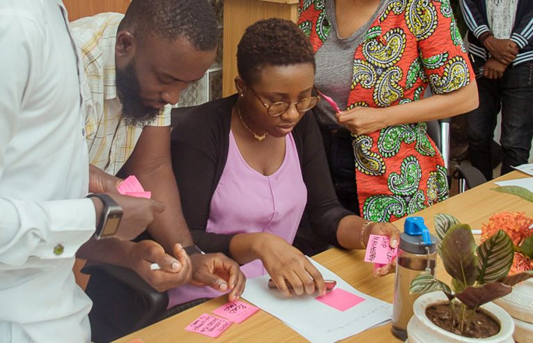 Photo of four participants. They're sitting and standing around a table and doing an activity together with sticky notes.