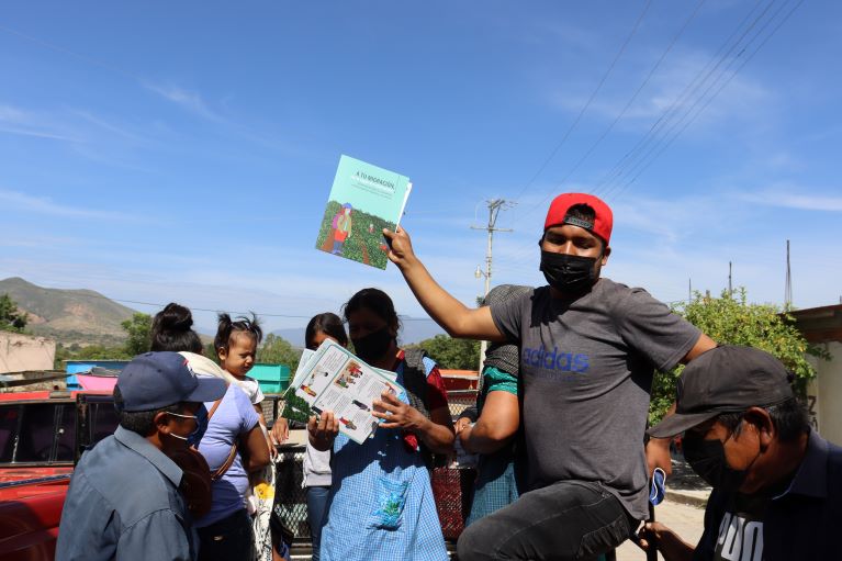 Photo of a man wearing a mask holding some materials about safer migration. He is surrounded by men, women and children with the same materials.