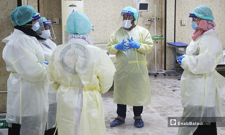 Photo of five medical workers who are talking in an emergency room. They're standing in a circle and wearing hairnets, face shields, face masks, gloves, and scrubs.