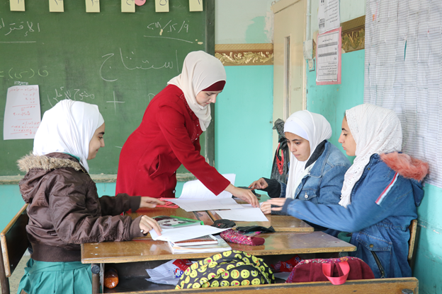 Photo of teacher standing at a table where 4 students are seated. The teacher is talking to one student while the other students listen.