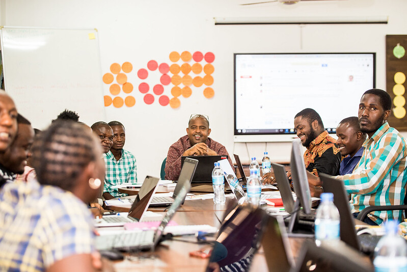 Photo of men and women sitting around a conference table with their laptops open, having a discussion. 