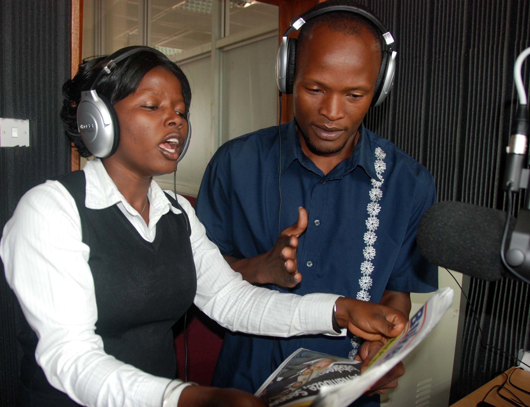 Photo of a male and female journalist in a radio studio, speaking into a microphone during a broadcast. 