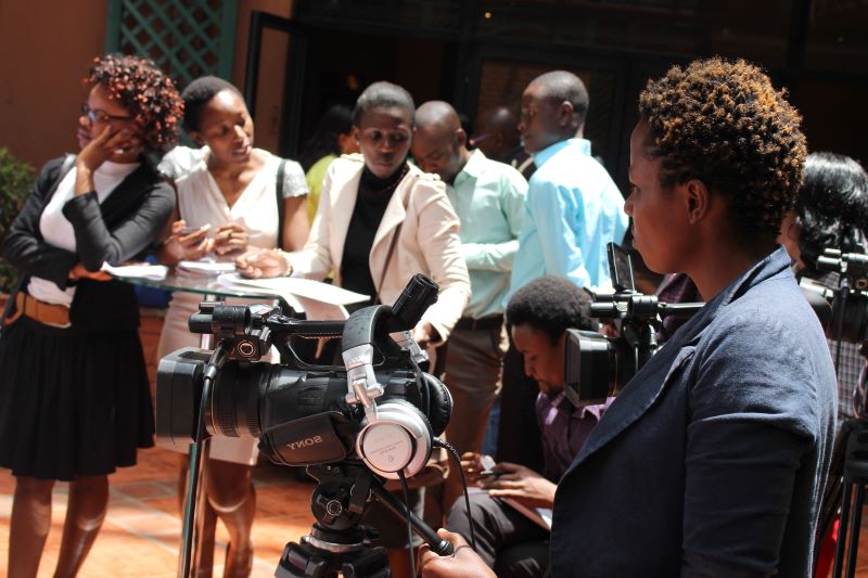 photo of journalists waiting for a press conference, standing behind their cameras and some are preparing to take notes. 