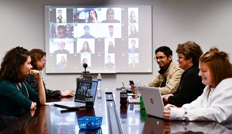 Group of students sitting at a table on laptops. Other participants in the background on a virtual call.