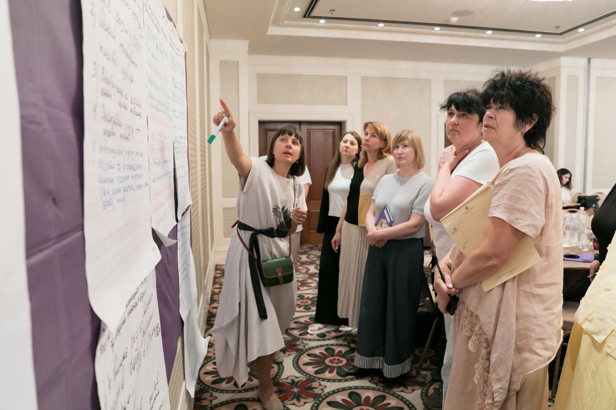 A group of six women standing in a conference room, attentively listening to one woman who is pointing at large notes on a wall.