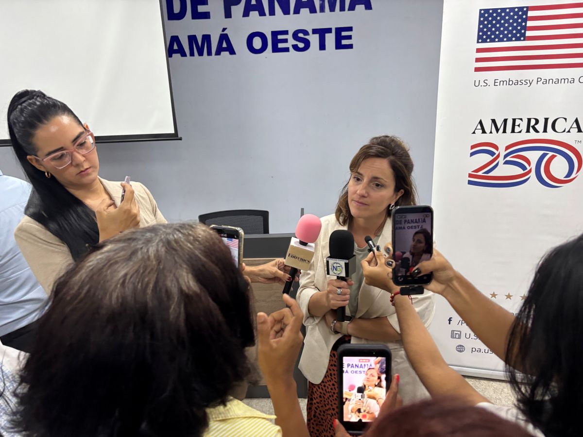 Laura Agosta speaking into multiple microphones held by reporters, with people recording her.