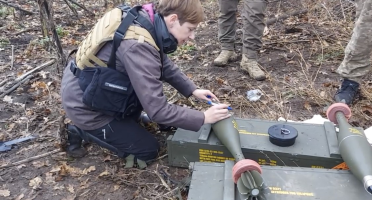 Picture of journalist in the field, kneeling next to ammunition, wearing personal protective equipment.
