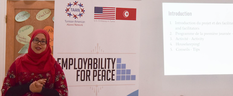 A young woman stands in front of a banner that reads Employability for Peace