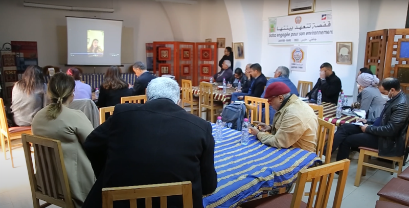 Photo of men and women sitting in a room at tables witching a presentation on a screen