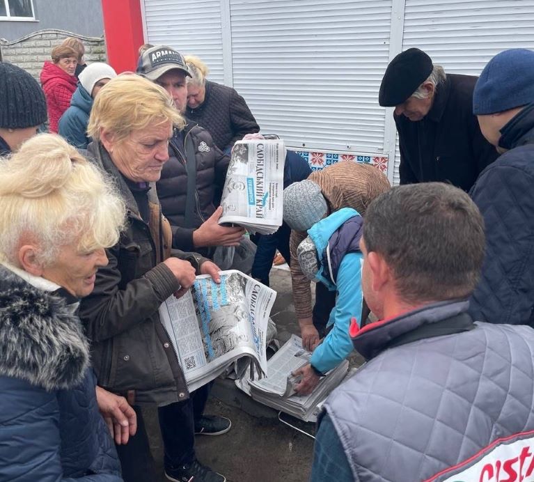 Photo of men and women crowding around a man distributing newspapers.