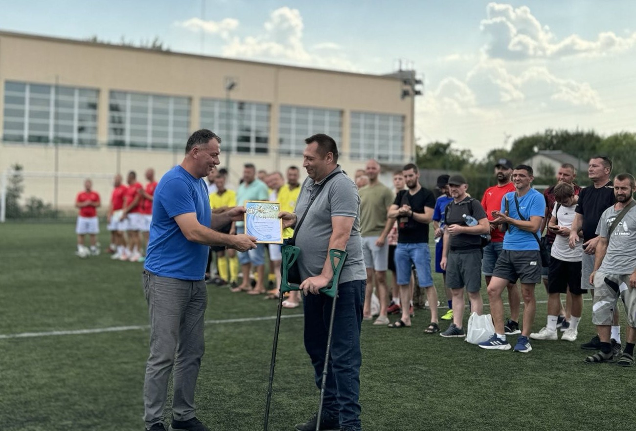 Photograph with one man handing a certificate to the other who uses crutches