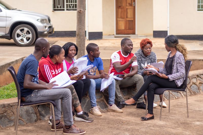 Male and female adult youth sitting outside a building discussing a paper. Two of the youth are sitting on chairs and the others are on a short wall. 