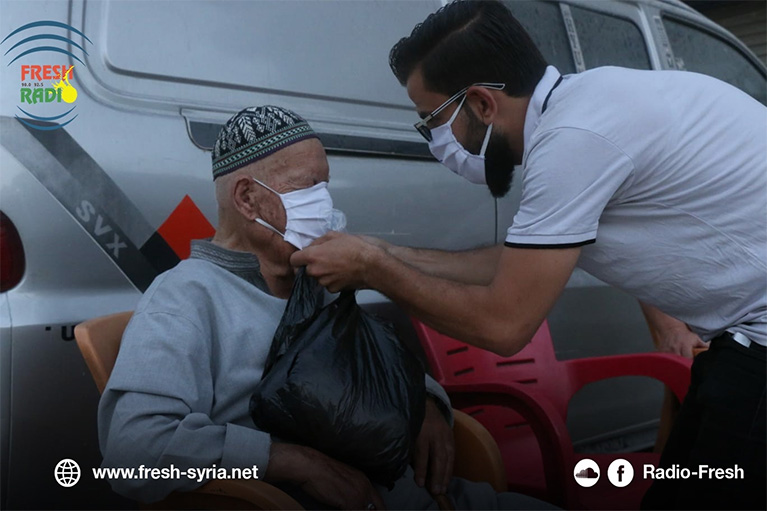 Photo of two people. One person is helping the other put on a face mask.