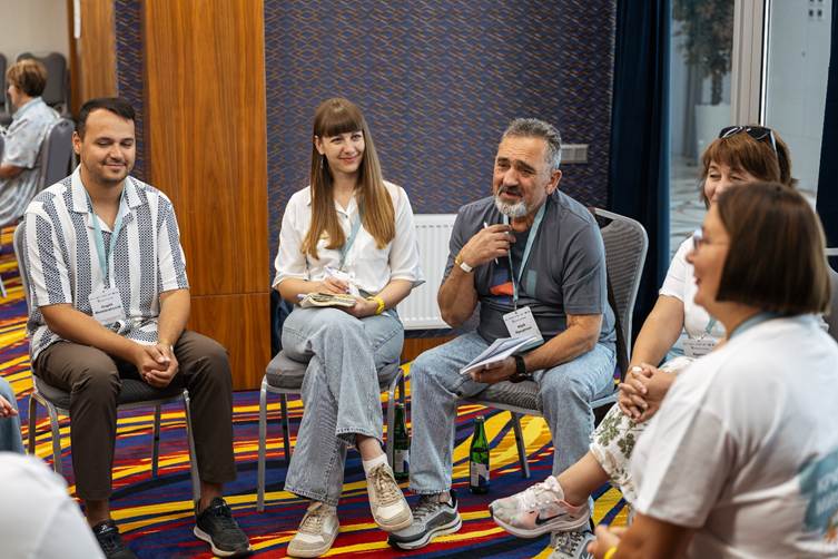 A group of people seated in a small conference discussion, with a bearded man speaking while holding notes