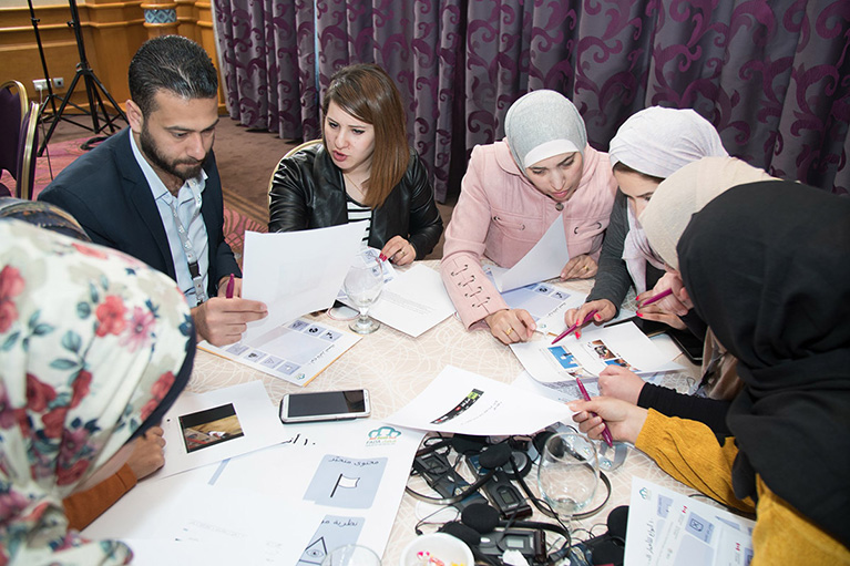 Photo of seven participants at a table in Jordan. They're looking at printouts and talking to each other.
