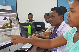 A woman pointing at a computer screen and talking to a colleague next to her while other participants work on laptops.