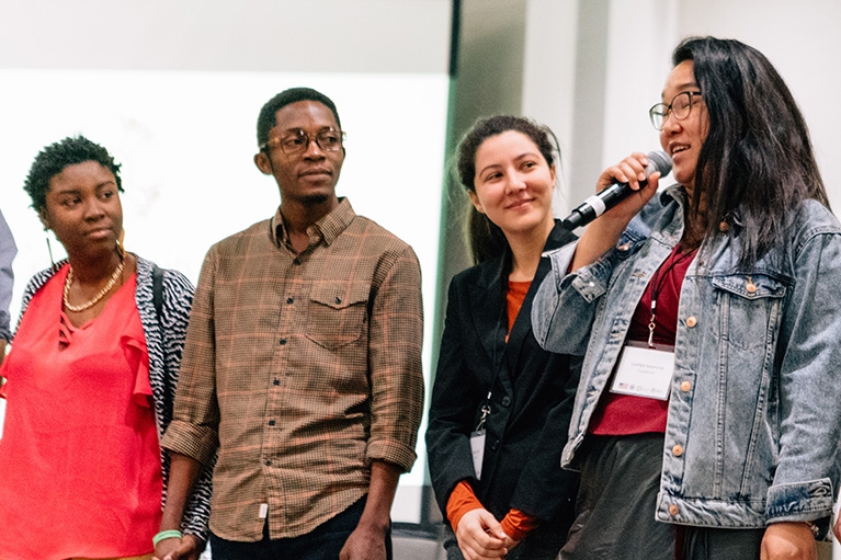 Photo of four people who are standing in a line. One person is holding a microphone and talking to the audience.