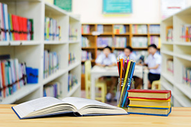 An open book, a jar of pencils, and a stack of books on a table in a library. In the background, three children are sitting around a table.