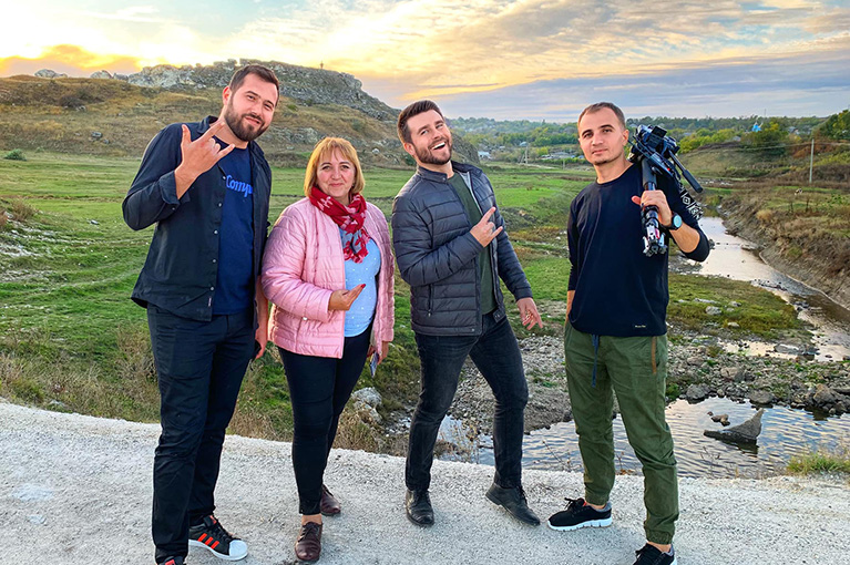 Photo of four people from the cast and crew of Tur-Retur. They're standing on a road in front of a hill and stream in Moldova.