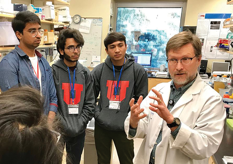 Four participants in the UGRAD-Pakistan program listen to a researcher in a lab coat at a university.