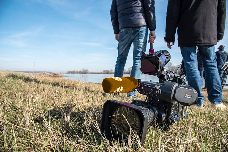 Photo of a video camera in the grass on an assignment. A reporter and crew are standing in the background and talking to each other.