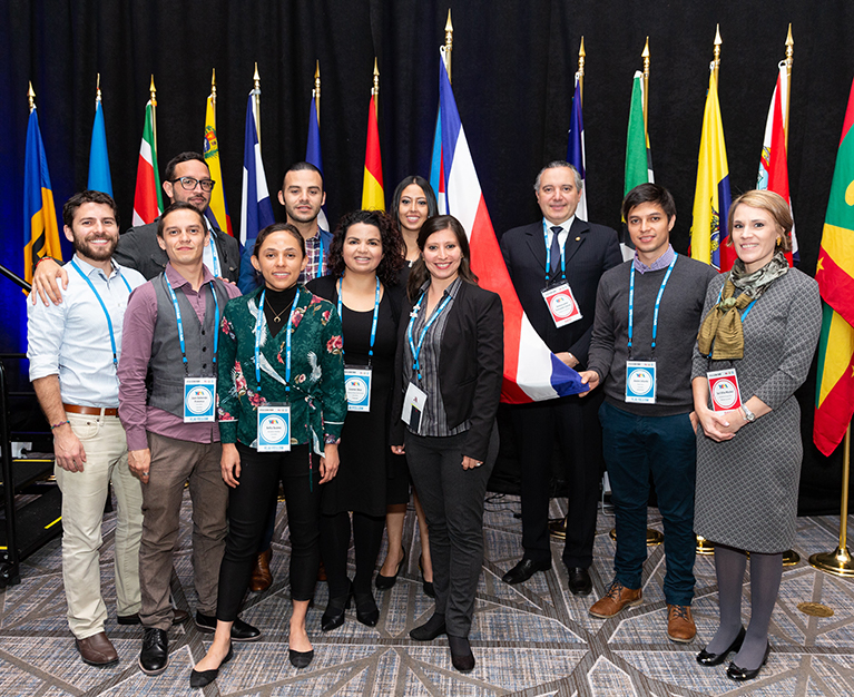 Photo of a group of YLAI participants in front of a dark curtain and thirteen flags.
