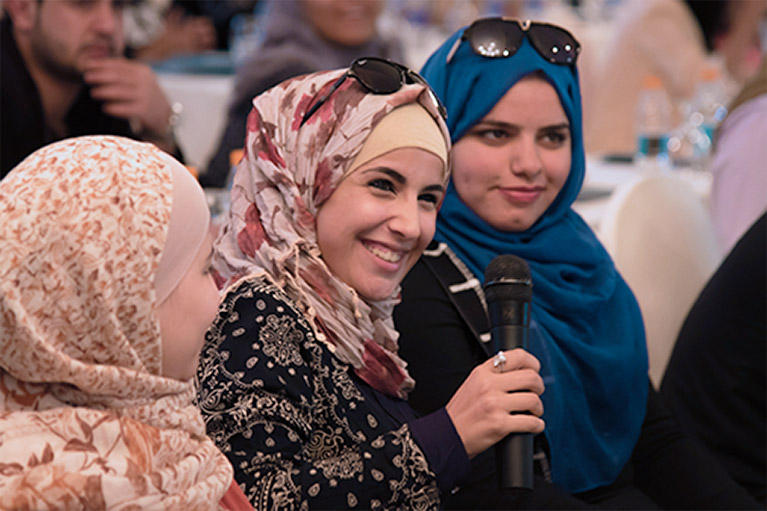 Three young women at an event in Jordan. One of the women is holding a microphone.