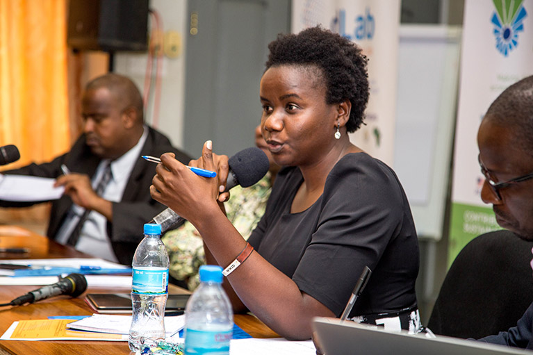 A woman speaking into a microphone at a meeting in Tanzania while two men take notes