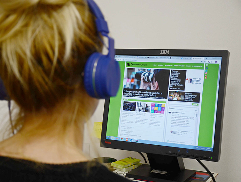 Photo of a person who is wearing headphones and looking at a news site on a desktop monitor.