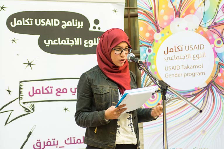 USAID Takamol participant standing in front of a microphone,  presenting information to an audience.