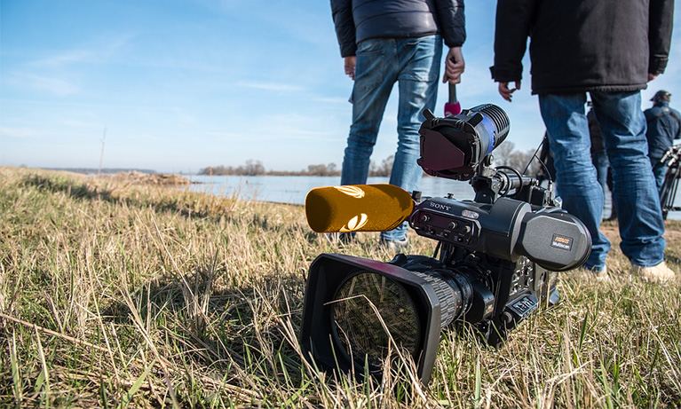 Photo of a video camera in the grass on an assignment. A reporter and crew are standing in the background and talking to each other.