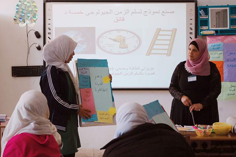 A teacher standing in the front of a classroom watching a student give a presentation.