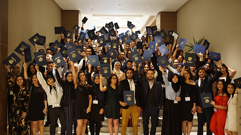 Dozens of participants from the TJSP program. They're standing on stairs in a hallway and holding up folders that show the U.S. Department of State's seal.