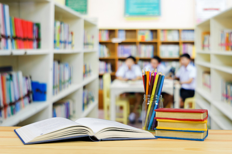 An open book, a jar of pencils, and a stack of books on a table in a library. In the background, three children are sitting around a table.