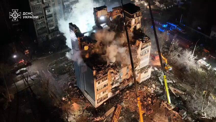 A damaged building with smoke rising, surrounded by emergency vehicles and cranes. 