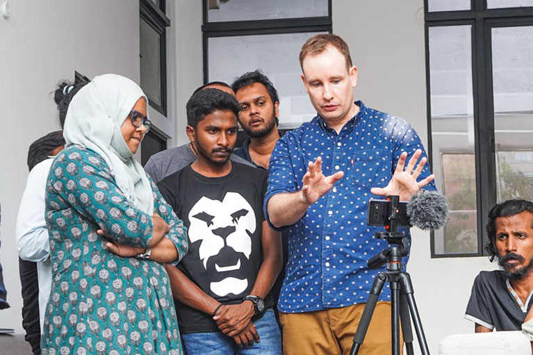 Participants and a facilitator in a mobile journalism workshop look at a video camera on a tripod.